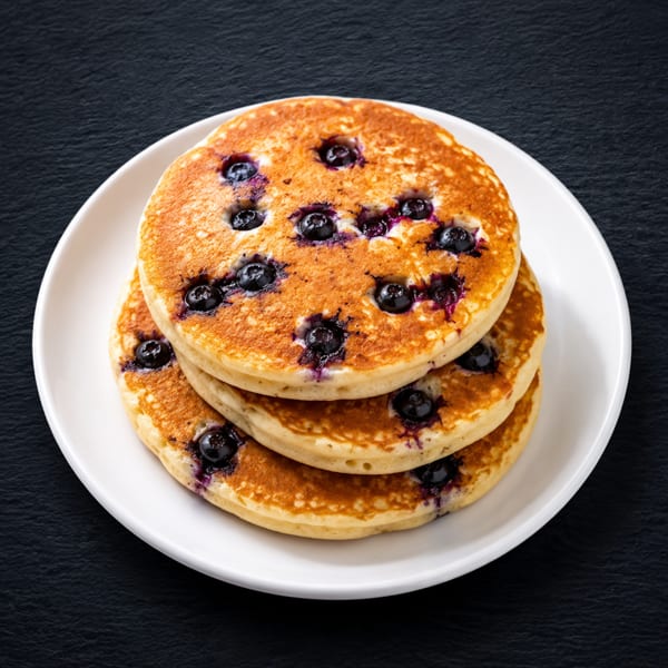 Stack of three golden blueberry pancakes on a white plate against a dark background.