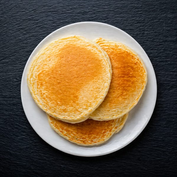 Three golden-brown pancakes stacked on a white plate, centered on a dark, textured surface.