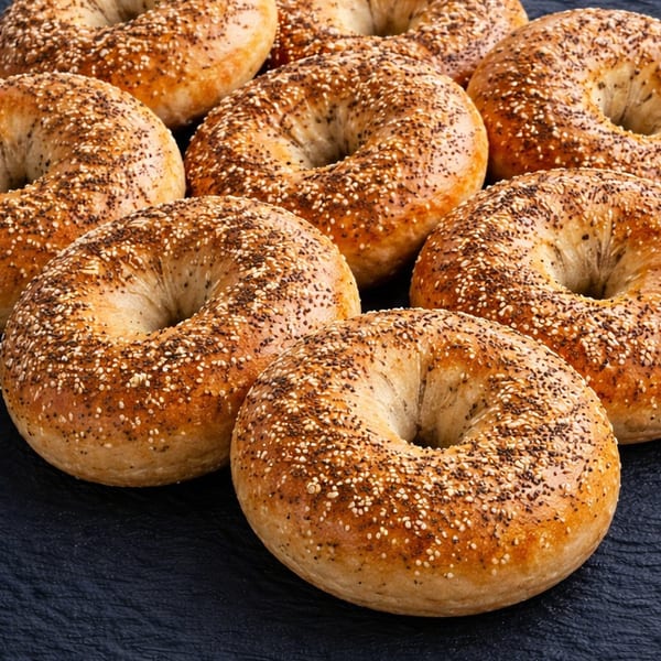 Several golden-brown everything bagels, generously topped with seeds, arranged on a dark surface.